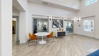Located at 1533 Ellis Rd in Durham, NC, this single-floor community space is a thoughtfully designed gathering hub that blends modern aesthetics with functional versatility. The open-plan layout seamlessly connects the kitchen and living room, creating a bright, welcoming environment bathed in natural light from large windows and double glass entry doors.

The living area commands attention with soaring ceilings, light wood-look flooring, and a striking feature wall clad in white shiplap, anchored by a wall-mounted flat-screen TV and bold navy blue cabinetry with gold hardware. Plush royal blue velvet seating and a patterned area rug add a vibrant, sophisticated touch. Recessed and built-in open shelving with warm wood accents provide display space that balances style and practicality.

The kitchen is equally impressive, showcasing rich navy cabinetry paired with crisp white quartz countertops and classic white subway tile backsplash. A stainless steel refrigerator, undermount sink, and a generous kitchen island with seating for multiple guests make this space ideal for entertaining. A separate dining nook with a round marble-top table and modern chairs overlooks glass-paneled windows into an adjacent fitness center, further enhancing the sense of openness and community throughout the space.