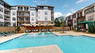 This amenity space is part of a contemporary multi-story residential community and is designed to support an active lifestyle. The outdoor pool area is a standout feature, showcasing a generously sized swimming pool with a shallow wading shelf lined with teal-cushioned lounge chairs. Surrounding the pool are multiple seating areas furnished with plush circular lounge pods and additional chaise lounges, all shaded by large umbrellas. Tiered retaining walls with decorative plantings, including palm trees, add a resort-like atmosphere to the space, set against the backdrop of the community's modern architecture.

The fitness center is equally impressive, occupying a bright, open interior space with large windows that allow ample natural light to fill the room. The flooring features a distinctive hexagonal patterned carpet throughout. Equipment includes treadmills, stationary bikes, a multi-functional cable and Smith machine, strength training machines, a full dumbbell rack, a punching bag, medicine balls, TRX suspension trainers, and a functional training rig. A large nature-themed mural adds a dynamic visual element, while pendant lighting and wall-mounted mirrors complete the polished, modern aesthetic of the space.