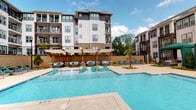 This amenity space is part of a contemporary multi-story residential community and is designed to support an active lifestyle. The outdoor pool area is a standout feature, showcasing a generously sized swimming pool with a shallow wading shelf lined with teal-cushioned lounge chairs. Surrounding the pool are multiple seating areas furnished with plush circular lounge pods and additional chaise lounges, all shaded by large umbrellas. Tiered retaining walls with decorative plantings, including palm trees, add a resort-like atmosphere to the space, set against the backdrop of the community's modern architecture.

The fitness center is equally impressive, occupying a bright, open interior space with large windows that allow ample natural light to fill the room. The flooring features a distinctive hexagonal patterned carpet throughout. Equipment includes treadmills, stationary bikes, a multi-functional cable and Smith machine, strength training machines, a full dumbbell rack, a punching bag, medicine balls, TRX suspension trainers, and a functional training rig. A large nature-themed mural adds a dynamic visual element, while pendant lighting and wall-mounted mirrors complete the polished, modern aesthetic of the space.