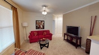 This single-floor living space is designed with comfort and functionality in mind. The living room serves as the centerpiece, featuring plush wall-to-wall carpeting in a neutral beige tone that adds warmth to the room. Crown molding runs along the perimeter of the ceiling, lending a classic, refined architectural touch throughout the space.

A ceiling fan with a light fixture provides both illumination and air circulation, complementing the room's practical design. The living area is spacious enough to accommodate a full-sized sofa, a round glass coffee table, and an entertainment setup, demonstrating a well-proportioned layout ideal for relaxing or entertaining.

A doorway visible in the background leads to an adjoining area with tile flooring, suggesting a transition to a kitchen, bathroom, or utility space. The neutral wall tones create a versatile backdrop that pairs well with a variety of décor styles.

Overall, the space offers a comfortable, move-in-ready feel with thoughtful architectural details, including clean lines, recessed lighting in the hallway, and a seamless flow between rooms that enhances the overall livability of the home.