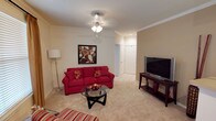 This single-floor living space is designed with comfort and functionality in mind. The living room serves as the centerpiece, featuring plush wall-to-wall carpeting in a neutral beige tone that adds warmth to the room. Crown molding runs along the perimeter of the ceiling, lending a classic, refined architectural touch throughout the space.

A ceiling fan with a light fixture provides both illumination and air circulation, complementing the room's practical design. The living area is spacious enough to accommodate a full-sized sofa, a round glass coffee table, and an entertainment setup, demonstrating a well-proportioned layout ideal for relaxing or entertaining.

A doorway visible in the background leads to an adjoining area with tile flooring, suggesting a transition to a kitchen, bathroom, or utility space. The neutral wall tones create a versatile backdrop that pairs well with a variety of décor styles.

Overall, the space offers a comfortable, move-in-ready feel with thoughtful architectural details, including clean lines, recessed lighting in the hallway, and a seamless flow between rooms that enhances the overall livability of the home.