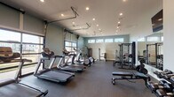 This fitness center is thoughtfully designed to support a comprehensive workout experience across two connected areas. The main exercise floor features durable rubber flooring and is lined with multiple treadmills and elliptical trainers positioned to take advantage of the expansive garage-style glass door along one wall, which floods the space with natural light and offers open outdoor views. Recessed ceiling lighting provides consistent illumination throughout. A strength training zone is outfitted with a power rack, adjustable bench, and a tiered dumbbell rack, giving users a full range of resistance training options. Wall-mounted mirrors in the strength area enhance the sense of space while allowing exercisers to monitor their form. A wall-mounted television is positioned for easy viewing. A dedicated spin studio is located in an adjoining room and features several cycling bikes arranged on rubber flooring beneath a mounted flat-screen television. Deep teal-toned walls and large windows provide a focused yet energizing atmosphere in this space. A rowing machine completes the cardio offerings. Together, both areas provide a versatile, well-lit environment suited for a wide variety of fitness routines.