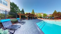 This outdoor amenity space centers around a generously sized swimming pool with clearly marked lane depths, its vivid blue water a striking focal point under open skies. The pool deck is finished with well-laid interlocking pavers, providing a durable and visually appealing surface throughout the area. A substantial wooden pergola structure serves as the centerpiece of the lounge zone, offering partial shade over a collection of reclining lounge chairs and small tables with cushioned seating in blue accents. Additional chaise lounges are positioned poolside, ideal for sunbathing. Classic-style lamp posts punctuate the space, suggesting the area is also functional during evening hours. The entire area is enclosed by decorative iron fencing, providing both security and a sense of defined space. Mature trees surrounding the perimeter offer natural screening and shade, enhancing privacy and creating a lush, resort-like atmosphere. The combination of structured architectural elements, open sky exposure, and greenery gives this outdoor space a well-balanced, inviting character suited for both relaxation and recreation.