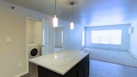 This single-floor residence is designed with an open-concept layout that seamlessly connects the kitchen and living areas. The kitchen serves as a central hub, anchored by a substantial island topped with speckled granite countertops and dark cabinetry beneath. Two cylindrical pendant lights hang above the island, providing focused task lighting while contributing to the space's modern aesthetic.

The open sightlines from the kitchen extend into the living area, which features light-toned carpeting and a large window that invites natural light into the space. Baseboard heating units are positioned along the walls, ensuring efficient climate control throughout. The neutral, light gray walls create a clean and versatile backdrop throughout the unit.

A dedicated laundry area is conveniently located just off the kitchen, housing a stacked or side-by-side washing unit, adding significant practical value to the home. Additional doorways visible from the main living space suggest access to bedrooms or other rooms beyond, all finished consistently with the same neutral tones. The overall ambiance is bright, functional, and move-in ready.