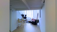 This two-floor space boasts a thoughtfully designed open-plan layout that seamlessly combines living and dining areas on the main level. Exposed wooden beams run across the white-painted ceiling, adding a distinctive architectural character and a sense of industrial charm to the interior. Large windows allow abundant natural light to pour in, enhancing the airy and spacious feel of the room.

The main living area features a casual seating arrangement with a red sofa and a dark leather armchair positioned around a low coffee table, set atop a patterned area rug over light hardwood flooring. A modular black shelving unit serves as both a functional storage solution and a design focal point, displaying decorative items. Adjacent to the living area, a compact dining setup includes a light wood high-top table paired with matching chairs, ideal for casual meals.

A staircase connects the main level to the upper floor, suggesting additional living or sleeping space above. The clean white walls throughout the space contribute to a minimal, contemporary aesthetic. Overall, this space offers a well-balanced blend of functionality and modern design, with architectural details and natural light as its standout features.