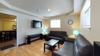 This single-floor space is thoughtfully designed for communal living, offering a welcoming and functional layout that balances comfort with practicality. The living area serves as the heart of the space, furnished with two dark leather sofas arranged around a low-profile coffee table, creating an inviting setting for relaxation and social interaction. A wall-mounted flat-screen TV and a sleek media console anchor one side of the room, while warm-toned ambient lamps and light hardwood flooring contribute to the bright, comfortable atmosphere. Multiple windows allow natural light to fill the lounge, enhancing its open and airy feel. Adjacent to the living area, the kitchen is well-appointed with stainless steel appliances, including an electric range with a ventilation hood, ample counter space with light-toned laminate surfaces, and wood-finish cabinetry offering generous storage. A utility sink and dedicated drying area add to the kitchen's practicality. The dining area features a dark high-top table with matching cross-back chairs, comfortably seating a group. Tile flooring in the kitchen and dining zone provides durability and easy maintenance. The overall design reflects a clean, modern aesthetic suited to shared occupancy, with neutral wall tones and efficient use of space throughout.