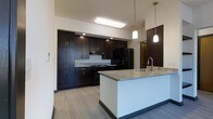 This single-floor space showcases a thoughtfully designed, contemporary kitchen as its centerpiece. Dark espresso-toned cabinetry lines the walls from floor to ceiling, providing generous storage while establishing a bold, modern aesthetic. The cabinetry is paired with light granite countertops that offer a striking contrast, adding texture and visual interest throughout the space.

A central kitchen island anchors the layout, topped with matching granite and equipped with a stainless steel sink. Pendant lighting hangs above the island, casting warm, focused light that enhances both functionality and ambiance. The main cooking area features a full suite of black appliances, including a gas range, built-in microwave, and a large refrigerator, all seamlessly integrated into the cabinetry design.

Recessed lighting and a flush ceiling light fixture provide bright, even illumination across the kitchen. The light hardwood-style flooring runs throughout, grounding the space with a clean, neutral foundation that complements the darker cabinetry tones. A doorway visible at the end of the kitchen corridor leads to an adjoining room, suggesting a well-connected and functional floor plan. Overall, the space delivers a polished, modern atmosphere suited for both everyday living and entertaining.