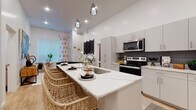 This well-appointed single-floor residence showcases a thoughtfully designed open-concept layout with modern finishes throughout. The kitchen serves as a centerpiece, featuring a generous quartz waterfall island with an integrated sink and seating for multiple bar-height wicker stools, sleek grey cabinetry, a dark tile backsplash, and a full suite of stainless steel appliances including a refrigerator, range, and over-the-range microwave. Pendant lighting and recessed fixtures illuminate the space, which flows naturally into the living area. The living room offers elevated ceilings, large windows dressed with decorative curtains and blinds that allow ample natural light, and warm light wood-style flooring that extends throughout the unit. A dedicated flex room provides versatility, currently configured with a desk workspace and daybed, ideal for a home office or guest space. The primary bedroom is a comfortable retreat with a window, recessed lighting, and warm-toned décor accents. Two full bathrooms complete the layout — one featuring a tub/shower combo with a large vanity mirror and white cabinetry, and the other offering a walk-in shower, open closet shelving, and light wood flooring. The overall ambiance is bright, clean, and contemporary.