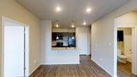 This single-floor space showcases a clean, modern design centered around an open-concept living and kitchen layout. The kitchen is defined by dark espresso cabinetry that contrasts sharply against light granite countertops, which extend across a peninsula that serves as both a workspace and casual dining surface. Black appliances, including a refrigerator and range, complement the cabinetry's bold tones. Two pendant lights hang above the peninsula, providing focused task lighting while adding a stylish touch to the space.

Recessed lighting throughout ensures consistent, well-distributed illumination across both the kitchen and adjoining living area. The living space features rich, dark hardwood-style flooring that runs continuously, grounding the open layout with warmth and visual continuity. Neutral beige walls keep the atmosphere light and versatile.

A doorway visible from the living area leads to an adjacent carpeted room, suggesting a clear separation between living and private spaces. The overall ambiance is contemporary and functional, with thoughtful design choices that balance practicality with aesthetic appeal, making the space feel cohesive and well-appointed throughout.