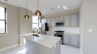 Located at 50 W Broad St in the heart of Columbus, Ohio, this single-floor space offers a refined, contemporary aesthetic highlighted by a thoughtfully designed kitchen. The kitchen serves as a central feature, showcasing soft gray cabinetry that extends to the ceiling, providing generous storage while maintaining a clean, cohesive look. A large central island with a bright white quartz countertop anchors the space, equipped with an undermount sink and a brushed gold faucet that adds a warm, modern accent. Overhead, two globe pendant lights in a gradient gold finish hang elegantly above the island, contributing both task lighting and visual interest. Stainless steel appliances, including a gas range, built-in microwave, and refrigerator, complement the kitchen's modern character. Recessed lighting throughout ensures the space is well-lit, while a window allows natural light to fill the area. The light-toned flooring ties the room together, reinforcing the bright, open ambiance. The overall design balances functionality with sophisticated style, making it a polished and practical space in a prime downtown location.