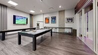 Located at 711 Treanor Avenue in Victoria, BC, this well-appointed games and recreation room offers a thoughtfully designed space for leisure and social activity. The room features contemporary finishes throughout, including durable wood-look laminate flooring, clean white walls, and a combination of recessed and pendant lighting that creates a bright, inviting atmosphere.

At the heart of the space sits a full-sized pool table with a sleek dark frame and grey felt surface, illuminated by a modern hanging light fixture. Alongside it, a regulation shuffleboard table provides additional entertainment, positioned beneath a large wall-mounted flat-screen television — ideal for watching sports while playing. A second large-screen TV is also mounted on an adjacent wall, ensuring excellent visibility from multiple areas of the room.

A built-in counter with bar-height stools along one wall serves as a casual spectator area, while framed sports memorabilia and artwork add personality and character to the décor. Decorative vertical slat accents and a concrete-effect feature wall introduce subtle textural contrast. A glass-paneled sliding door provides access to an adjoining area, enhancing the open and connected feel of the space. Natural light filters in through windows, complementing the well-distributed overhead lighting for a vibrant, energetic ambiance.