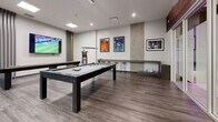 Located at 711 Treanor Avenue in Victoria, BC, this well-appointed games and recreation room offers a thoughtfully designed space for leisure and social activity. The room features contemporary finishes throughout, including durable wood-look laminate flooring, clean white walls, and a combination of recessed and pendant lighting that creates a bright, inviting atmosphere.

At the heart of the space sits a full-sized pool table with a sleek dark frame and grey felt surface, illuminated by a modern hanging light fixture. Alongside it, a regulation shuffleboard table provides additional entertainment, positioned beneath a large wall-mounted flat-screen television — ideal for watching sports while playing. A second large-screen TV is also mounted on an adjacent wall, ensuring excellent visibility from multiple areas of the room.

A built-in counter with bar-height stools along one wall serves as a casual spectator area, while framed sports memorabilia and artwork add personality and character to the décor. Decorative vertical slat accents and a concrete-effect feature wall introduce subtle textural contrast. A glass-paneled sliding door provides access to an adjoining area, enhancing the open and connected feel of the space. Natural light filters in through windows, complementing the well-distributed overhead lighting for a vibrant, energetic ambiance.