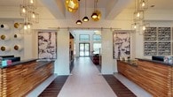 This single-floor clubhouse is a meticulously designed amenity space that blends contemporary aesthetics with eclectic, artistic details. The reception area makes an immediate impression, featuring dual wood-and-stone counters flanked by large abstract artworks, sliding glass barn doors on brass hardware, and a striking cluster of geometric gold and matte pendant lights suspended from soaring ceilings. Mixed tile and wood-plank flooring guides visitors through the space.

Beyond the entry, a lounge area commands attention with a dramatic dark fireplace wall adorned with a longhorn skull mount, a tiered bookshelf display, acrylic accent chairs, and a sculptural multi-bulb chandelier. Natural light pours in through floor-to-ceiling windows and clerestory panels, enhancing the airy volume of the room.

A dedicated entertainment nook features undulating wood-paneled walls and ceiling, plush velvet seating, globe pendant lights, and built-in display screens, creating an intimate, immersive atmosphere. Adjacent to this is a game room with a pool table, curated moon-phase artwork, and views to the outdoors.

The kitchenette is outfitted with dark granite countertops, warm wood cabinetry, white upper cabinets, a mosaic tile backsplash, and open industrial shelving. A welcoming entry vestibule with skylights and a package area rounds out this well-appointed community facility.