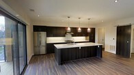 This contemporary single-floor space showcases a thoughtfully designed open-concept kitchen that serves as the centerpiece of the layout. The kitchen is outfitted with rich, dark espresso cabinetry that extends from floor to ceiling, providing generous storage while creating a bold visual contrast against the crisp white countertops and backsplash. A spacious central island offers ample workspace and seating potential, finished with the same clean white surface that ties the design together. Three elegant glass pendant lights hang above the island, adding a warm, refined touch to the space. Stainless steel appliances, including a full-size French door refrigerator and a built-in range with an overhead hood, complete the high-end kitchen ensemble. Recessed ceiling lighting supplements the pendant fixtures, ensuring the space is well-lit throughout. The warm-toned hardwood flooring runs continuously through the open area, lending a sense of warmth and cohesion to the modern aesthetic. The clean lines, neutral palette, and quality finishes throughout reflect a contemporary design sensibility, making this a functional and visually striking space suited for both everyday living and entertaining.