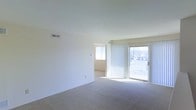 This single-floor residential space offers a clean, functional layout filled with natural light. The main living area is generously proportioned, featuring wall-to-wall neutral-toned carpeting that adds warmth and comfort underfoot. Crisp white walls keep the space feeling fresh and open, providing a versatile backdrop for a variety of interior styles.

A standout feature of the living area is the full-height sliding glass door dressed with vertical blinds, which floods the room with daylight and provides direct access to an outdoor balcony or patio area. Through the glass, views of the surrounding exterior are visible, offering a connection to the outdoors. An additional window in the adjacent room further enhances the natural light throughout the space.

The layout flows naturally from the living area into a connecting room, suggesting a well-organized floor plan designed for everyday practicality. Ceiling-mounted HVAC vents are present throughout, ensuring consistent climate control year-round.

Overall, the space presents a bright and airy atmosphere with a simple, neutral aesthetic that lends itself well to personalization. Its open feel, ample natural light, and accessible outdoor connection make it a comfortable and practical living environment.