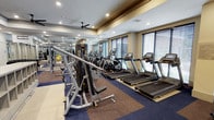 This fitness center offers a comprehensive workout environment designed to accommodate a wide range of exercise needs. The space is organized into distinct functional zones, with cardio equipment — including multiple treadmills and elliptical machines — positioned along a windowed wall fitted with horizontal blinds that allow natural light to filter in while maintaining privacy. The strength training area features a variety of cable and weight machines arranged to maximize usable floor space.

The interior design reflects a polished, hotel-style aesthetic, with warm beige walls accented by a crown molding detail along the upper perimeter and a tray ceiling element that adds architectural depth. Recessed lighting throughout ensures consistent illumination across the entire space, complemented by a ceiling fan that promotes air circulation. Full-length mirrors line one wall, enhancing the sense of openness and allowing users to monitor their form during workouts. Durable, multi-toned carpet tiles cover the floor in the strength training zone, while a lighter hard-surface flooring defines the cardio area. The overall ambiance is clean, functional, and well-maintained.