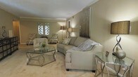 This single-floor space offers a thoughtfully designed interior centered around a welcoming living area. The room is finished in soft, warm neutral tones — from the taupe-painted walls to the plush, light-colored carpeting — creating a cohesive and calming atmosphere throughout. Crown molding adds a refined architectural detail along the ceiling line, lending a classic elegance to the space.

The living area is furnished with a curved, upholstered sofa accented by decorative throw pillows, complemented by a pair of patterned armchairs that add visual texture without disrupting the room's understated palette. A glass-and-metal coffee table serves as a stylish focal point at the center of the seating arrangement, while additional glass side tables reinforce the space's contemporary sensibility.

Ambient lighting plays a key role in the room's character, with floor lamps and table lamps casting a warm, inviting glow. A striking wall art panel with a vertical, reflective design anchors one wall, adding a modern sculptural element to the space. Decorative lattice-style window treatments allow soft light to filter in while maintaining a sense of privacy. Overall, the space presents a harmonious blend of classic architectural details and contemporary furnishings, resulting in a polished and comfortable environment.