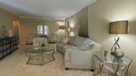 This single-floor space offers a thoughtfully designed interior centered around a welcoming living area. The room is finished in soft, warm neutral tones — from the taupe-painted walls to the plush, light-colored carpeting — creating a cohesive and calming atmosphere throughout. Crown molding adds a refined architectural detail along the ceiling line, lending a classic elegance to the space.

The living area is furnished with a curved, upholstered sofa accented by decorative throw pillows, complemented by a pair of patterned armchairs that add visual texture without disrupting the room's understated palette. A glass-and-metal coffee table serves as a stylish focal point at the center of the seating arrangement, while additional glass side tables reinforce the space's contemporary sensibility.

Ambient lighting plays a key role in the room's character, with floor lamps and table lamps casting a warm, inviting glow. A striking wall art panel with a vertical, reflective design anchors one wall, adding a modern sculptural element to the space. Decorative lattice-style window treatments allow soft light to filter in while maintaining a sense of privacy. Overall, the space presents a harmonious blend of classic architectural details and contemporary furnishings, resulting in a polished and comfortable environment.