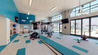 This well-appointed fitness center is designed to accommodate a wide range of workout routines within a single, expansive single-story space. The interior is immediately distinguished by its soaring ceilings and floor-to-ceiling windows along one side, which flood the facility with natural light and provide open views of an outdoor pool area and surrounding landscape. Large glass doors offer direct access to the exterior, further connecting the indoor and outdoor environments.

The design scheme is modern and energetic, featuring teal textured wall panels paired with crisp white walls and ceilings. The flooring consists of a patterned carpet tile in neutral tones accented by teal blocks, which visually define workout zones throughout the space. Multiple wall-mounted flat-screen televisions are positioned throughout the gym for convenience.

The fitness floor is comprehensively equipped, featuring cardio machines including treadmills, ellipticals, and stationary bikes, as well as a multi-station cable and strength machine setup, a punching bag, free weights, dumbbells, medicine balls, and battle ropes. A separate glass-enclosed room serves as a dedicated studio space for cycling or group fitness. Large ceiling fans provide additional airflow, and restroom access is available directly within the facility.