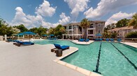This two-story residential community features an impressive resort-style outdoor amenity area designed for relaxation and recreation. At the heart of the complex lies a generously sized leisure pool with a shallow entry and a dedicated lap pool equipped with clearly marked lanes, both maintained with crystal-clear water. The expansive pool deck is outfitted with an abundance of blue chaise lounge chairs and large market umbrellas, providing ample space for sunbathing and socializing. A standout feature of the outdoor area is the collection of elegant private cabanas, each fitted with canopied daybed loungers that offer a shaded retreat for residents. A pergola-style seating structure adjacent to the pools adds an additional covered gathering space. The surrounding landscape is enriched by mature palm trees and lush greenery, creating a tropical ambiance throughout. A well-appointed clubhouse with covered colonnaded porches sits poolside, seamlessly connecting indoor and outdoor living. The multi-story residential buildings feature private balconies that overlook the pool area, reinforcing the community's open and airy character. The entire pool complex is enclosed by a secure perimeter fence, ensuring privacy and safety for all residents.