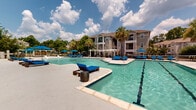 This two-story residential community features an impressive resort-style outdoor amenity area designed for relaxation and recreation. At the heart of the complex lies a generously sized leisure pool with a shallow entry and a dedicated lap pool equipped with clearly marked lanes, both maintained with crystal-clear water. The expansive pool deck is outfitted with an abundance of blue chaise lounge chairs and large market umbrellas, providing ample space for sunbathing and socializing. A standout feature of the outdoor area is the collection of elegant private cabanas, each fitted with canopied daybed loungers that offer a shaded retreat for residents. A pergola-style seating structure adjacent to the pools adds an additional covered gathering space. The surrounding landscape is enriched by mature palm trees and lush greenery, creating a tropical ambiance throughout. A well-appointed clubhouse with covered colonnaded porches sits poolside, seamlessly connecting indoor and outdoor living. The multi-story residential buildings feature private balconies that overlook the pool area, reinforcing the community's open and airy character. The entire pool complex is enclosed by a secure perimeter fence, ensuring privacy and safety for all residents.