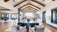 Located at 300 Intergraph Way in Madison, Alabama, this impressively appointed single-floor clubhouse offers a generous and thoughtfully designed interior. The centerpiece of the space is the expansive open-concept living area, distinguished by dramatic vaulted ceilings adorned with rich, dark-stained exposed wood beams and a stylish chandelier. Floor-to-ceiling windows flood the room with natural light and provide sweeping views of the outdoor pool area, while a striking stone fireplace column with an integrated TV serves as a bold architectural focal point. Warm hardwood flooring runs throughout, complementing the neutral palette and upscale furnishings, including tufted leather sofas and a dark stone coffee table. The adjacent dining area features large sliding glass doors that open directly to the pool terrace, enhancing indoor-outdoor connectivity. The kitchen is well-equipped and visually striking, with white shaker cabinetry, dark stone countertops, a large island, stainless steel appliances, and a vibrant green subway tile backsplash. A separate office provides a functional workspace with built-in desks, ample storage, and a small seating area. Together, these spaces create a polished, welcoming environment suited for both relaxation and community gathering.
