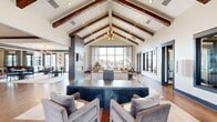 Located at 300 Intergraph Way in Madison, Alabama, this impressively appointed single-floor clubhouse offers a generous and thoughtfully designed interior. The centerpiece of the space is the expansive open-concept living area, distinguished by dramatic vaulted ceilings adorned with rich, dark-stained exposed wood beams and a stylish chandelier. Floor-to-ceiling windows flood the room with natural light and provide sweeping views of the outdoor pool area, while a striking stone fireplace column with an integrated TV serves as a bold architectural focal point. Warm hardwood flooring runs throughout, complementing the neutral palette and upscale furnishings, including tufted leather sofas and a dark stone coffee table. The adjacent dining area features large sliding glass doors that open directly to the pool terrace, enhancing indoor-outdoor connectivity. The kitchen is well-equipped and visually striking, with white shaker cabinetry, dark stone countertops, a large island, stainless steel appliances, and a vibrant green subway tile backsplash. A separate office provides a functional workspace with built-in desks, ample storage, and a small seating area. Together, these spaces create a polished, welcoming environment suited for both relaxation and community gathering.