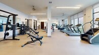 Located at 1703 Rockhill Rd in McKinney, TX, this well-appointed fitness center offers a generously sized, single-level layout designed to accommodate a comprehensive range of workout activities. The space is characterized by its bright, modern aesthetic, featuring crisp white walls, light-toned flooring, and high ceilings fitted with ceiling fans and linear LED lighting that contribute to an open, energizing atmosphere.

The main workout area is lined with floor-to-ceiling windows and roller shades, flooding the interior with abundant natural light and offering views of the surrounding outdoor environment. A row of treadmills and elliptical machines lines the windowed wall, creating an inviting cardio zone. Adjacent to this, a strength training section features a multi-station cable machine, free weights with a dedicated dumbbell rack, and adjustable benches.

Structural columns divide the space naturally, while large wall-mounted mirrors enhance the sense of depth and allow users to monitor their form. A separate functional training area, framed by sleek black-framed glass doors, houses a rowing machine, a Pilates-style bench, and additional specialty equipment, providing a versatile zone for varied training modalities. Built-in wooden shelving and an adjacent utility area round out the practical amenities of this well-equipped facility.