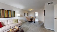 This single-floor residence offers a thoughtfully arranged layout that balances comfort and functionality throughout. The living room serves as the heart of the home, featuring plush wall-to-wall carpeting, neutral gray walls, and ample seating including a sofa and armchair. A vibrant multicolored artwork anchors the space, while a window with blinds allows for natural light and ventilation. The living area flows seamlessly into a dedicated dining space, where a round glass-top table surrounded by four cushioned chairs creates an inviting setting for meals. A wall-mounted mirror and closet add practicality to the dining area. The kitchen is equipped with rich dark wood cabinetry, light granite countertops, a classic white subway tile backsplash, and stainless steel appliances including a gas range, dishwasher, refrigerator, and over-the-range microwave, along with light wood-style flooring. The bedroom is a relaxing retreat with carpeted floors, a ceiling fan, natural light from a window with blinds, and ample wall space. The full bathroom features light blue tile flooring, a tub-shower combination with textured wall tile, a dark wood vanity, and a well-lit mirror. Overall, the space presents a clean, cohesive aesthetic with warm neutral tones throughout.