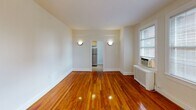 Located at 829 Quincy St NW in Washington, DC's Petworth neighborhood, this second-floor apartment offers a well-organized single-level layout with classic character and practical finishes throughout. The living area is a standout space, showcasing richly toned, polished hardwood floors that extend through the main living spaces and bedroom. Soft wall sconces provide warm ambient lighting, while multiple windows fitted with blinds allow natural light to fill the rooms and offer views of the surrounding neighborhood. The bedroom mirrors the living area's aesthetic with the same gleaming hardwood floors, a ceiling light fixture, and a window-mounted air conditioning unit alongside a radiator, ensuring year-round comfort. The kitchen is equipped with light wood cabinetry offering ample storage, a dark stone-style countertop, a stainless steel refrigerator, and a gas range, all set on durable tile flooring. The bathroom features a full-size soaking tub and shower combination with clean white tile surround and chrome fixtures, along with overhead cabinetry for additional storage. The layout flows efficiently from room to room, making practical use of the available space while maintaining a cohesive, bright, and neutral aesthetic throughout.