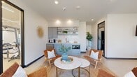 This thoughtfully designed single-floor space combines functionality and modern aesthetics in a compact, efficient layout. The kitchenette serves as a focal point, featuring soft gray-blue lower cabinetry, open floating shelves, a subway tile backsplash, a built-in sink, microwave, and under-counter appliances — all finished with clean white countertops. Adjacent to the kitchenette, the open lounge area is anchored by a round marble-top coffee table with gold legs, surrounded by woven leather accent chairs set atop a natural jute rug, creating a warm and inviting gathering space. A decorative sunburst wall accent and potted fiddle-leaf fig tree add organic, artistic touches throughout. A dedicated office nook is set apart from the lounge, featuring a light wood desk, rounded upholstered guest chairs, a wall-mounted display screen, and a softly textured accent wall with abstract wallcovering. Polished concrete flooring flows seamlessly throughout the space, lending an urban, contemporary feel. Floor-to-ceiling black-framed glass doors and windows at the entrance allow generous natural light to pour in while offering views of the outdoor surroundings, making the space feel open and connected to the exterior environment.