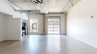 Located at 1741 36th St in Denver, CO, this single-floor space showcases a distinctly industrial aesthetic that blends raw architectural elements with modern functionality. Polished concrete floors stretch throughout the open layout, reflecting natural light and lending a clean, contemporary feel to the interior.

The main area is defined by soaring exposed concrete ceilings with visible ductwork, conduit, and structural elements that reinforce the space's loft-like character. A large glass-panel garage-style door serves as a dramatic architectural feature, flooding the interior with natural light and offering views of the surrounding neighborhood. A standard window further enhances the brightness of the space.

The kitchen is well-appointed with dark cabinetry, black granite countertops, open wall shelving, and a full suite of stainless steel appliances including a range, dishwasher, microwave, and refrigerator. A secondary utility area features navy blue cabinetry with sinks and open shelving, providing additional workspace and storage.

A clean, tiled bathroom includes a walk-in shower and modern fixtures. Additional open rooms with white walls and exposed overhead piping offer flexible, adaptable space suitable for a variety of uses. The overall ambiance is modern and utilitarian, ideal for creative, professional, or live-work configurations.
