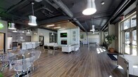 This expansive single-floor amenity lounge showcases a striking blend of industrial and contemporary design. The soaring dark-painted ceiling features exposed ductwork and mechanical systems, lending the space an urban, loft-inspired character. Statement pendant lights hang at varying heights, casting a warm, inviting glow across the rich hardwood flooring that runs throughout.

At the heart of the space is a central feature wall housing a sleek fireplace topped by a wall-mounted flat-screen television, framed by curated wall art. Surrounding this focal point is a generously sized seating area outfitted with modern white sofas, accent chairs, and coffee tables — ideal for casual gatherings or quiet lounging. A warm wood slat ceiling detail above this zone adds a refined contrast to the industrial aesthetic overhead.

Toward one end of the space, a bar-height counter with white stools provides a dedicated spot for socializing, complemented by what appears to be a café or beverage station. On the opposite side, a billiards table anchors a game area, adding a recreational dimension to the lounge. Large windows along the perimeter draw in abundant natural light, contributing to the open, airy atmosphere of this thoughtfully designed communal space.