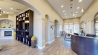 This single-story community facility is defined by its refined Mediterranean-inspired architecture, characterized by a series of elegant arched doorways, soaring vaulted ceilings, and warm neutral tones throughout. Wood-look tile flooring flows consistently across the space, lending a cohesive and polished aesthetic.

The reception area serves as the focal point of the main entrance, anchored by a curved, dark-finished reception desk beneath pendant globe lighting. Recessed lighting and tall ceilings amplify the openness of this area. A built-in dark wood shelving unit displays decorative accents, while arched niches punctuate the walls. A workstation area with desks and seating is positioned near large windows, allowing ample natural light and outdoor views.

Adjacent lounge areas offer comfortable seating, including tufted sofas and accent chairs in blue and yellow tones, complemented by contemporary wall art. A cozy nook with a pass-through window provides an intimate seating spot.

The fitness center is generously proportioned with towering ceilings, featuring a full suite of cardio equipment, cable machines, free weights, and exercise mats. Large glass partitions connect the gym visually to a game room equipped with a billiards table, chandelier lighting, and additional seating.