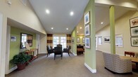 This single-story space presents a well-appointed common lounge area designed for comfort and social interaction. The room features vaulted ceilings that enhance the sense of openness, complemented by recessed lighting that provides consistent, warm illumination throughout. Large windows dressed with striped curtains allow natural light to fill the space, creating an inviting atmosphere.

The seating area is thoughtfully arranged with upholstered armchairs positioned to encourage conversation, centered on a neutral carpet that anchors the grouping. The walls are finished in a calming sage green and warm beige palette, adding visual interest while maintaining a relaxed tone.

A gallery-style wall display along the green accent column features a series of framed landscape artworks, adding a decorative focal point to the space. Additional framed artwork is distributed across the walls, reinforcing the curated, hospitable character of the interior.

Adjacent areas are visible through open passageways, suggesting the lounge connects to additional functional spaces. A small accent table along one wall adds a practical touch. Overall, the space balances functionality with a warm, welcoming aesthetic suited to communal use.