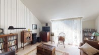 This single-floor residence offers a well-organized layout that balances comfort and character throughout its rooms. The living area is a standout space, featuring vaulted ceilings with distinctive vertical wood panel accent walls and large sliding glass doors fitted with vertical blinds that flood the room with abundant natural light. Soft carpeting and an eclectic mix of vintage furnishings lend the space a warm, lived-in feel.

The adjacent dining area connects seamlessly to the kitchen via a breakfast bar with barstool seating, creating an open and sociable flow. The kitchen is well-equipped with ample white cabinetry, generous counter space, a double-basin sink, and tile flooring.

The primary bedroom continues the vaulted ceiling aesthetic, offering generous proportions, a window with natural light, and plush carpet underfoot. A walk-in closet provides notable storage. The second bedroom also benefits from vaulted ceilings and a window, offering flexibility as a sleeping or multipurpose room.

The bathroom features a wide vanity with a large mirror, white cabinetry, and tile flooring. A carpeted hallway connects the private rooms efficiently. Throughout the residence, the recurring vaulted ceilings and natural light create an airy, spacious ambiance complemented by warm, classic detailing.