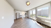 This well-designed one-bedroom, one-bathroom unit offers a thoughtful layout that maximizes its compact footprint. The open-concept main living area seamlessly connects the kitchen and dining space, featuring light hardwood-style flooring, neutral grey walls, and a flush ceiling light fixture. The modern kitchen is outfitted with white flat-panel cabinetry, a white subway tile backsplash, stainless steel appliances including a refrigerator, electric range, microwave, and dishwasher, complemented by track lighting overhead. Large floor-to-ceiling windows flood the living area with abundant natural light while offering open views of the surrounding cityscape and greenery beyond — a standout feature of this unit. A long, clean hallway connects the main living space to the private bedroom and bathroom. The bedroom is carpeted for comfort, with soft neutral tones and a sliding closet. The full bathroom is finished with a white vanity featuring an integrated countertop sink, a combination tub and shower, wood-look tile flooring, and a large mirror. Throughout the unit, the design aesthetic is consistently clean, modern, and functional, creating a bright and practical living environment.