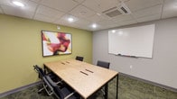 This single-floor professional meeting space is designed to support focused collaboration and productive discussions. At the heart of the room sits a light natural wood conference table with built-in cable management slots, surrounded by ergonomic black rolling chairs that comfortably accommodate a small group. The room features a combination of green-accented and neutral grey walls, lending a modern yet approachable atmosphere. A large whiteboard mounted on the grey wall provides ample space for brainstorming and presentations, complete with a marker tray for convenience. Adding a touch of creativity and visual interest, a vibrant abstract artwork adorns the green accent wall, bringing warmth and energy to the space. Overhead, a drop ceiling with recessed lighting and a ceiling-mounted HVAC unit ensures even illumination and a comfortable climate throughout. The patterned carpeted flooring ties the room together with a subtle professional aesthetic. This well-appointed conference room strikes a balance between functionality and style, making it well-suited for team meetings, training sessions, and collaborative work sessions.
