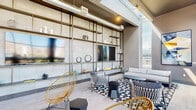This contemporary amenity lounge is defined by its striking industrial-modern aesthetic, combining soaring ceilings with exposed ductwork, skylight panels, and an expansive floor-to-ceiling window wall that floods the interior with natural light. The space is anchored by a dramatic full-wall shelving unit constructed of metal and stone tile, housing a large flat-screen television, curated décor objects, and display shelving — creating a bold, gallery-like focal point.

The main lounge area features an eclectic mix of seating, including a neutral toned sofa, woven rattan accent chairs in warm gold tones, and geometric side tables atop a black-and-white patterned area rug. A secondary seating nook hosts a trio of framed botanical and landscape artworks above additional gold accent chairs, offering a quieter, more intimate setting.

Along the window wall, a sleek counter-height bar runs the length of the glass, lined with teal and black woven bar stools. This workspace-style seating area frames sweeping views of the surrounding cityscape and mountain backdrop, creating an inspiring environment for work or socializing. Light wood-toned flooring ties the areas together seamlessly, while curated artwork and thoughtful lighting throughout contribute to the space's polished, urban character.
