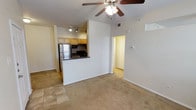 This single-floor space showcases an open-concept design that seamlessly connects the kitchen and living area. The living space features vaulted ceilings that lend an airy, spacious feel, complemented by a ceiling fan with integrated lighting. Neutral-toned carpet covers the living area, while tile flooring defines the kitchen transition zone.

The kitchen is positioned as a semi-open peninsula layout, partially separated from the living area by a raised breakfast bar with a dark countertop. Light wood cabinetry lines the kitchen walls, providing ample storage, while a track lighting system illuminates the workspace. Stainless steel appliances, including a full-size refrigerator, and black appliances complete the kitchen setup.

A hallway visible from the living area leads to additional rooms, suggesting a practical and functional floor plan. The walls throughout are finished in a clean, neutral white, contributing to a bright and cohesive atmosphere. A thermostat is wall-mounted near the hallway entrance, and a wall vent ensures consistent climate control throughout the space. Overall, the layout balances openness with defined functional zones.
