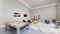 Located at 8011 Augusta Pines Dr in Spring, TX, this single-floor recreational lounge is a well-appointed game and social space designed for community entertainment. The room immediately impresses with its soaring tray ceilings and recessed lighting, creating an open, airy atmosphere throughout the generously sized interior.

The space is anchored by a full-size pool table positioned prominently near the center, complemented by a foosball table and a shuffleboard table along the perimeter — offering a well-rounded selection of classic game room entertainment. Multiple wall-mounted flat-screen televisions are thoughtfully positioned around the room, ensuring clear sightlines from various seating areas.

Comfortable lounge seating, including a large sectional reclining sofa and additional armchairs, are arranged to encourage relaxed socializing. A bistro-style high-top table with bar stools provides an additional casual gathering spot. The flooring features a bold, modern patterned carpet in shades of gray, yellow-green, and white, adding a lively visual element to the neutral gray walls.

Natural light filters in through multiple windows fitted with blinds, offering views of the surrounding greenery. French-style glass doors connect to adjacent rooms, enhancing the open flow of the space. Decorative wall sconces add a subtle stylistic touch, rounding out this versatile and inviting recreational environment.