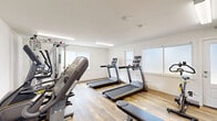 Located at 2490 Lancaster Dr in Richmond, CA, this fitness center offers a well-equipped exercise space designed to support a variety of workout routines. The room features clean white walls, durable wood-look flooring, and bright fluorescent lighting that keeps the space feeling open and energized. Natural light filters in through frosted windows along the walls, adding a pleasant brightness to the interior.

The exercise room is outfitted with a comprehensive selection of fitness equipment. Cardio options include multiple treadmills and a stationary spin bike, as well as an elliptical trainer for low-impact workouts. A Hoist multi-functional cable machine provides strength training versatility, while an adjustable workout bench supports a range of exercises. A Matrix elliptical trainer rounds out the cardio offerings, giving users multiple options for aerobic conditioning.

The layout is thoughtfully arranged to maximize the available floor area, allowing users to move between equipment with ease. Safety features include a mounted fire extinguisher and clearly marked exit signage. Fitness center rules are posted on the wall, ensuring a structured and safe environment for all users. The space offers a practical and well-maintained setting for residents seeking a convenient on-site fitness facility.