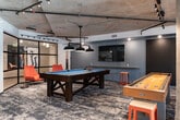 Located in Auburn, Washington, this single-floor amenity space blends industrial design with bold, colorful interiors to create a dynamic and inviting environment. Exposed concrete ceilings with visible ductwork and track lighting lend an urban, loft-like character to the space, while large windows invite natural light and views of the surrounding area.

The main lounge area features a generous sectional sofa in slate blue, accented with mustard and teal throw pillows, anchored by a patterned area rug in deep blue and grey tones. A curated gallery of vibrant, eclectic artwork lines the walls, adding personality and visual energy throughout.

Adjacent to the lounge is a dedicated game room, enclosed with sleek black-framed glass partitions. This area is equipped with a full-size pool table, a shuffleboard table, and casual orange accent seating, all beneath an industrial exposed-ceiling with pendant lighting.

A well-appointed kitchenette serves the space with light wood cabinetry, a matte black faucet, built-in appliances, and open shelving displaying colorful decorative vessels. A cozy nook with a textured brick-style feature wall, built-in bench seating in a vibrant multicolored fabric, and a round café table provides an intimate spot for smaller gatherings. Together, these areas form a cohesive, amenity-rich space ideal for socializing and recreation.
