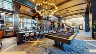 Located at 5180 Peachtree Blvd in Chamblee, Georgia, this generously sized amenity lounge delivers a sophisticated, resort-style experience across a single sprawling floor. The space is defined by soaring double-height ceilings, exposed wood beam accents, and dramatic arched architectural details that lend a grand, club-like atmosphere. Large floor-to-ceiling glass doors flood the interior with natural light while offering views to the exterior.

The main lounge area features bold geometric large-format tile flooring and is anchored by multiple seating arrangements, including high-back wingback chairs, leather sofas, and conversation groupings set atop richly patterned area rugs. A striking sculptural chandelier with exposed Edison bulbs serves as a centerpiece overhead.

A long bar with dark countertops and stylish bar seating is equipped with large display screens, making it ideal for social gatherings. Adjacent to the bar, a billiards table and foosball and ping pong tables provide ample entertainment options. Floor-to-ceiling dark-toned bookshelves with decorative displays line the walls, adding warmth and depth to the open layout.

A dedicated private poker room offers an intimate setting with a round poker table, leather chairs, built-in illuminated shelving, and a wall-mounted flat-screen TV. Throughout the space, curated artwork and layered lighting create a polished, inviting ambiance suited for both relaxation and socializing.