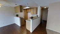 This single-floor space is designed with practicality and functionality in mind. The kitchen serves as the focal point, showcasing an abundance of light wood cabinetry that provides generous storage both above and below the countertops. The layout features a peninsula-style configuration with two breakfast bar extensions, offering additional counter space and a natural divider between the kitchen and the adjoining living area. The countertops display a granite-style surface that adds a touch of durability and subtle elegance to the workspace. A built-in overhead light fixture illuminates the main cooking and prep area, ensuring the kitchen remains bright and well-lit. The flooring throughout is rich, dark hardwood, which creates a striking contrast against the white walls and light cabinetry, lending the space a warm yet contemporary feel. A textured popcorn ceiling adds a classic residential character overhead. HVAC vents are integrated into the ceiling, ensuring climate comfort throughout. A hallway visible from the kitchen leads further into the residence, hinting at additional rooms beyond. Overall, the space balances a clean, neutral aesthetic with practical design elements suited for everyday living.