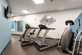 Located at 140 Mayhill St in Saddle Brook, NJ, this well-equipped fitness center offers a comprehensive workout environment across a single floor. The space is organized into distinct functional zones, each designed to support a variety of exercise routines. The main cardio area features a row of TRUE treadmills positioned along a motivational "FITNESS" word-art wall mural, creating an energizing atmosphere for endurance training. A separate room with steel-blue accent walls houses elliptical trainers and stationary cycling bikes, separated from the broader facility by elegant French-style glass-panel doors. The strength training section is outfitted with Paramount multi-station cable machines and free weight equipment, complemented by large wall-mounted mirrors that enhance the sense of space and allow users to monitor their form. Natural light filters into the strength area through frosted windows. Durable speckled rubber flooring runs throughout, providing both safety and resilience underfoot. A connecting corridor lined with mirrors leads to restroom and utility facilities, keeping the layout practical and user-friendly. The overall design balances functionality with a clean, modern aesthetic, making this fitness center a capable and inviting space for residents or members looking to maintain an active lifestyle.