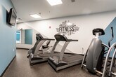 Located at 140 Mayhill St in Saddle Brook, NJ, this well-equipped fitness center offers a comprehensive workout environment across a single floor. The space is organized into distinct functional zones, each designed to support a variety of exercise routines. The main cardio area features a row of TRUE treadmills positioned along a motivational "FITNESS" word-art wall mural, creating an energizing atmosphere for endurance training. A separate room with steel-blue accent walls houses elliptical trainers and stationary cycling bikes, separated from the broader facility by elegant French-style glass-panel doors. The strength training section is outfitted with Paramount multi-station cable machines and free weight equipment, complemented by large wall-mounted mirrors that enhance the sense of space and allow users to monitor their form. Natural light filters into the strength area through frosted windows. Durable speckled rubber flooring runs throughout, providing both safety and resilience underfoot. A connecting corridor lined with mirrors leads to restroom and utility facilities, keeping the layout practical and user-friendly. The overall design balances functionality with a clean, modern aesthetic, making this fitness center a capable and inviting space for residents or members looking to maintain an active lifestyle.