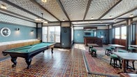 This single-floor space is a thoughtfully curated social lounge that draws heavily from vintage pub design traditions. The interior is immediately distinguished by its boldly patterned wallpaper and matching ceiling treatments, featuring intricate botanical and geometric motifs framed by dark wood beams that add architectural definition throughout. The vivid, multicolored encaustic tile flooring anchors the space, blending geometric star patterns in warm reds, blues, and yellows, complemented by layered area rugs.

A carved, dark wood bar with an ornate mirrored back panel serves as a central focal point, reflecting craftsmanship typical of classic saloon design. Adjacent to the bar, a full-size billiards table with a green felt surface occupies a generous portion of the space, illuminated by a pendant light fixture overhead. Seating areas include tufted leather banquette seating paired with dark wood tables, as well as bistro-style tables surrounded by green upholstered stools.

Natural light filters in through multiple sets of tall wood-framed windows and French doors that lead to an outdoor terrace with a railing, offering open skyline views. A wall-mounted television and framed artwork contribute to the lounge's layered, character-rich ambiance.