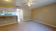This single-story space is designed with a practical, straightforward layout that connects its key living areas seamlessly. The living room is finished with neutral-toned carpet and warm beige walls, creating a welcoming atmosphere. A ceiling fan with a built-in light fixture is centrally mounted on the textured ceiling, providing both airflow and illumination. The space opens toward the kitchen and hallway, lending an open, connected feel to the floor plan.

The kitchen is well-equipped with white cabinetry above and below the counter, offering ample storage space. Wood-toned countertops pair with dark hardwood-style flooring, and appliances include a white electric coil range, a dishwasher, and a stainless steel sink positioned beneath upper cabinets. A range hood is installed above the stove for ventilation.

The bedroom is generously sized, featuring the same neutral carpet and beige walls found throughout the unit. A large window dressed with vertical blinds allows natural light to fill the room, casting bright patterns across the floor. A louvered closet door provides access to a built-in closet with shelving. A ceiling fan is also present, maintaining consistent comfort across the space.
