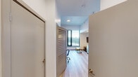 This thoughtfully designed single-floor unit offers a clean, contemporary aesthetic throughout, with light wood-style flooring and crisp white walls that create a bright and cohesive atmosphere. The open layout maximizes the available footprint, making efficient use of every square foot.

The kitchen is a standout feature, outfitted with a full suite of stainless steel appliances including an electric range, built-in microwave, dishwasher, and full-size refrigerator. Light wood cabinetry extends to the ceiling, providing ample storage, while white countertops offer a clean, modern contrast. Recessed lighting illuminates the workspace effectively.

The living area is anchored by a built-in Murphy bed with integrated shelving and cabinetry, allowing the space to function as both a comfortable lounge and a sleeping area. A sofa sits beneath the fold-down bed, and a large floor-to-ceiling window floods the room with natural light while offering elevated views of the surrounding cityscape.

The bathroom features a full tub-shower combo with tile surround, a toilet, and a sliding door entry, maintaining the unit's streamlined design. A dedicated entry area includes louvered closet doors for additional storage. The unit is equipped with a wall-mounted air conditioning unit, ensuring year-round comfort. Overall, the space is modern, functional, and well-appointed.